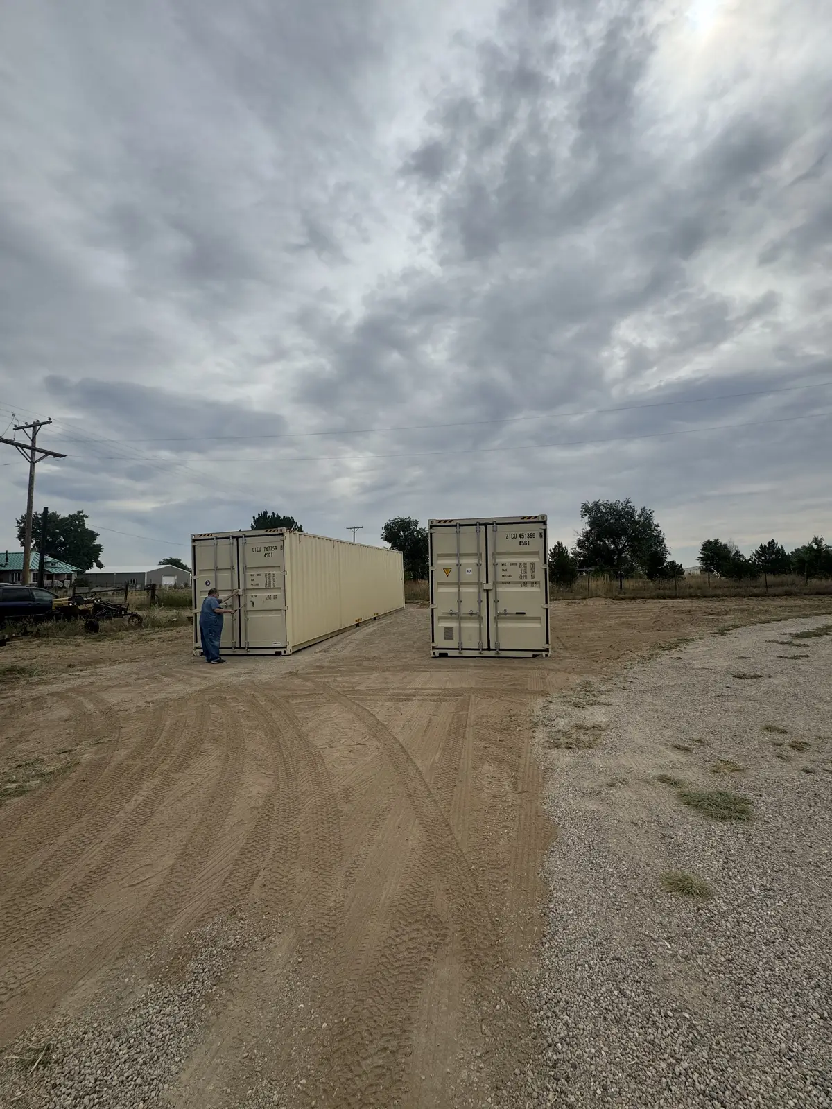 Two tan shipping containers on a dirt lot with a worker nearby