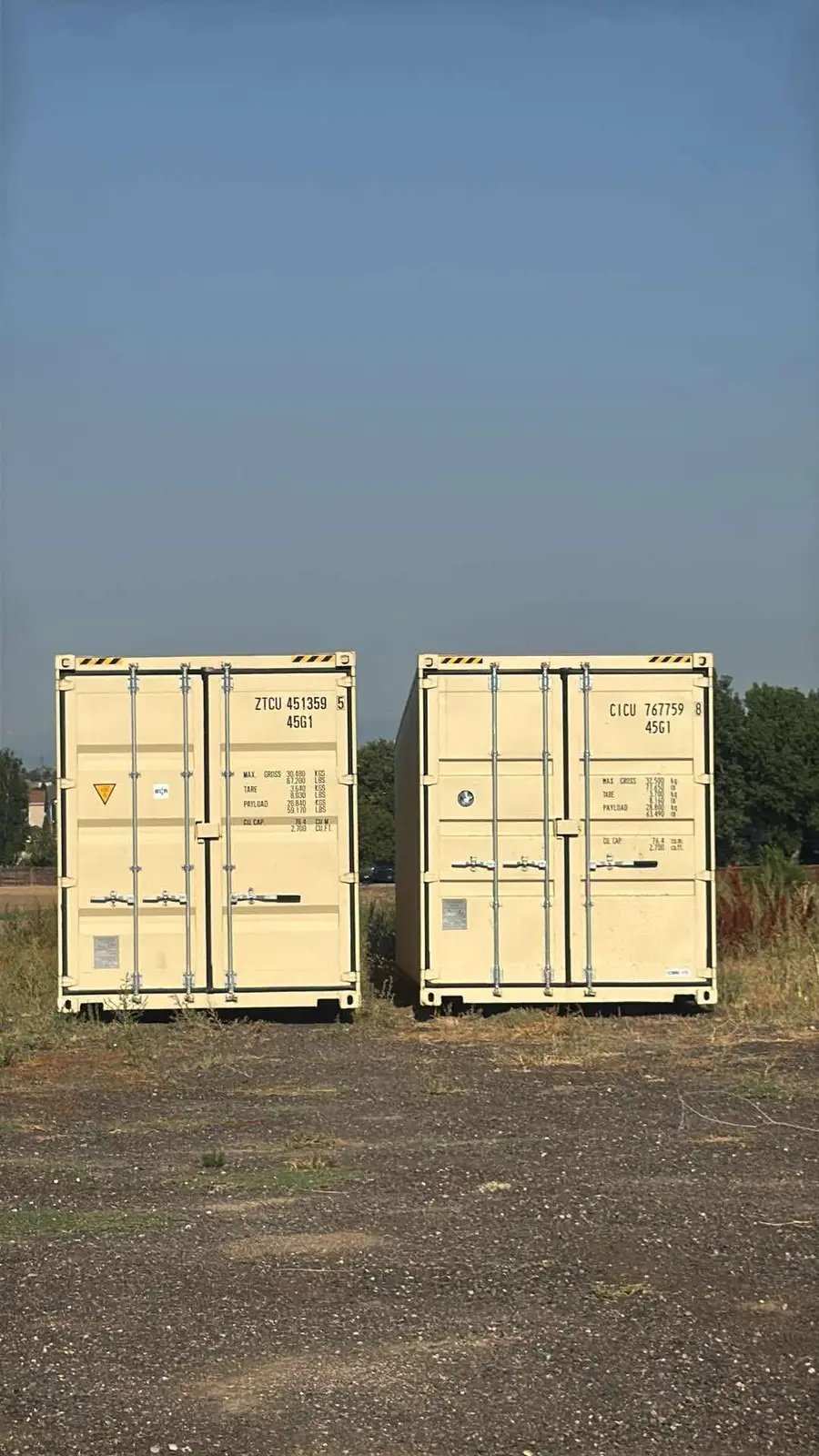 Two tan shipping containers side by side on an open rural lot