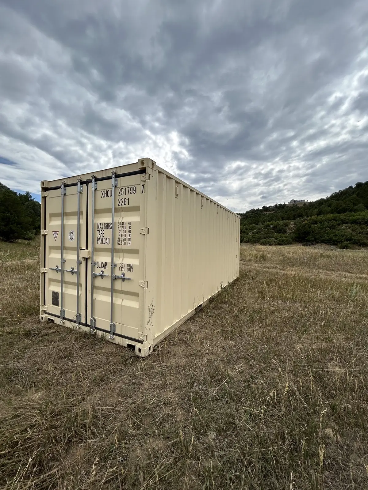 Tan shipping container sitting in a grassy field under a cloudy sky